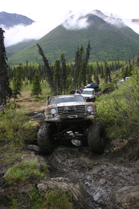 2003 Jeep TJ - Several off-road vehicles are seen traversing a challenging muddy and rocky path through a forested mountain landscape.