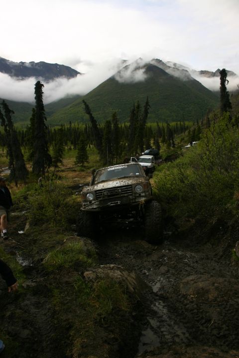 2003 Jeep TJ - Several off-road vehicles traverse a challenging muddy track amidst a backdrop of misty mountains and dense forest.
