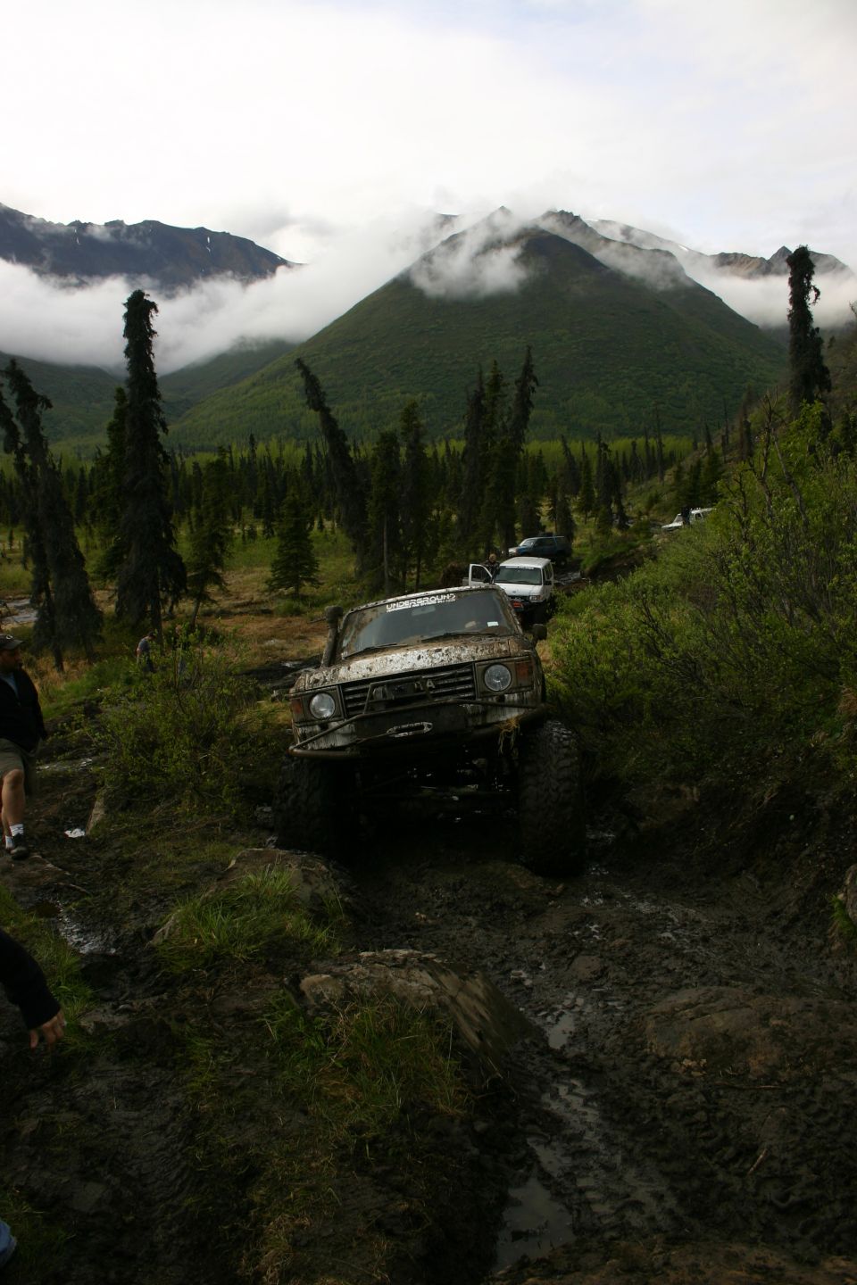 Mud-covered off-road vehicle navigating a muddy trail, with other vehicles behind it and a misty mountain landscape in the background