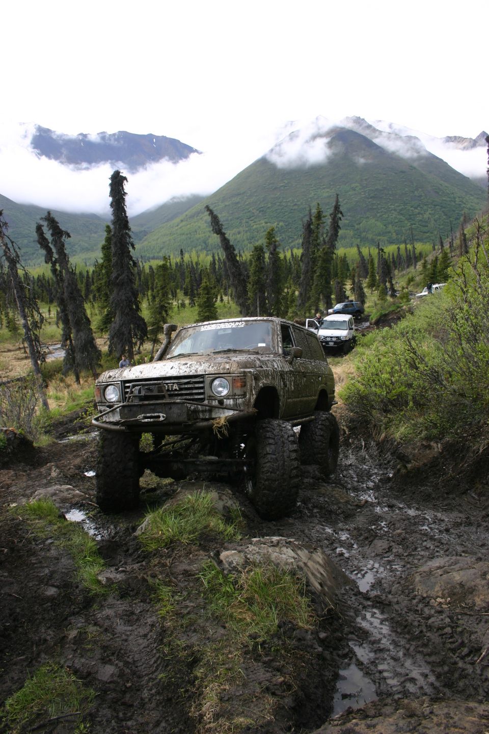 Muddy 1980 Toyota Land Cruiser FJ60 with snorkel and winch, driving on a dirt trail with other vehicles behind