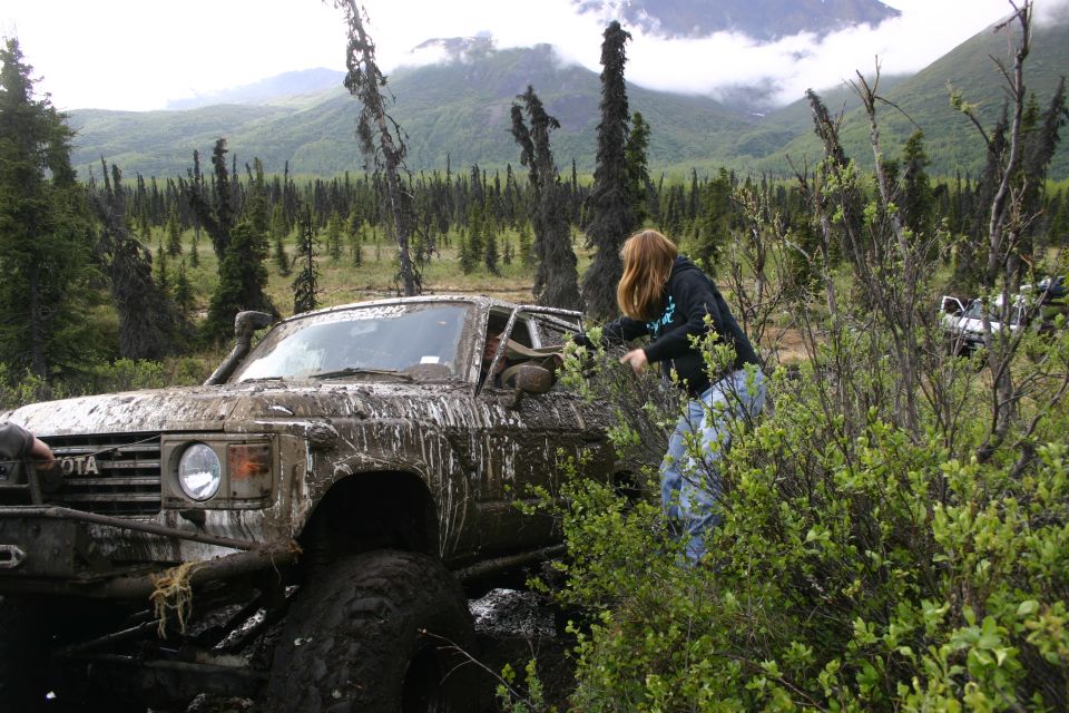 Muddy Toyota Land Cruiser stuck in brush with a person reaching in. Another white vehicle is visible in the background