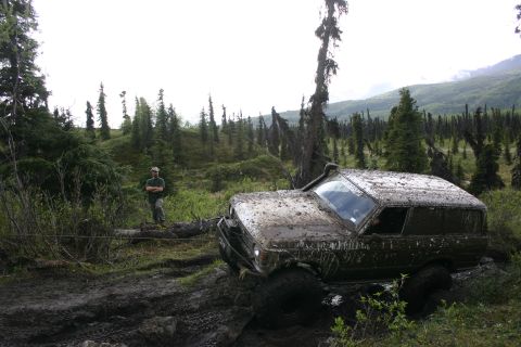 2003 Jeep TJ - A mud-covered SUV is stuck in a ditch while a person watches nearby in a wooded, mountainous area.