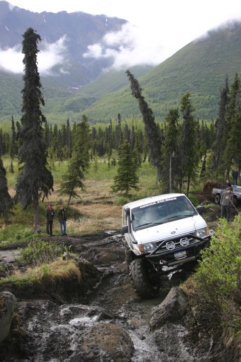 2003 Jeep TJ - An off-road vehicle navigates a challenging muddy terrain surrounded by dense forest and misty mountains.