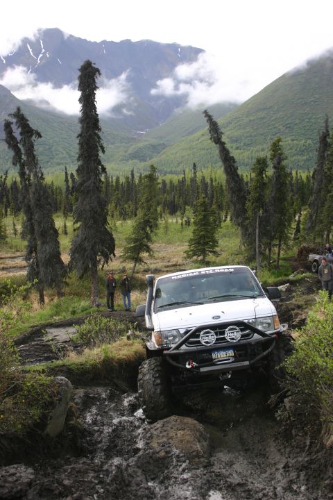 2003 Jeep TJ - A white off-road SUV tackles a challenging muddy track in a mountainous, forested area.