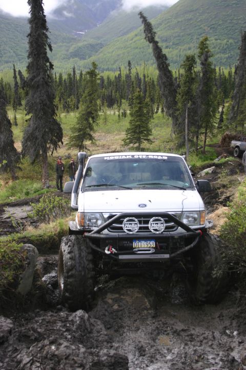 2003 Jeep TJ - A white van with a snorkel and custom off-road bumper navigates a muddy trail in a wooded area.