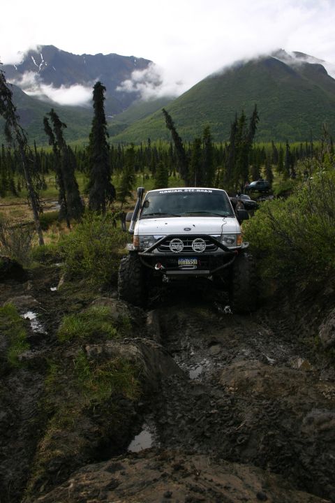 2003 Jeep TJ - Off-road SUV tackles a challenging muddy path in a mountainous, forested area.