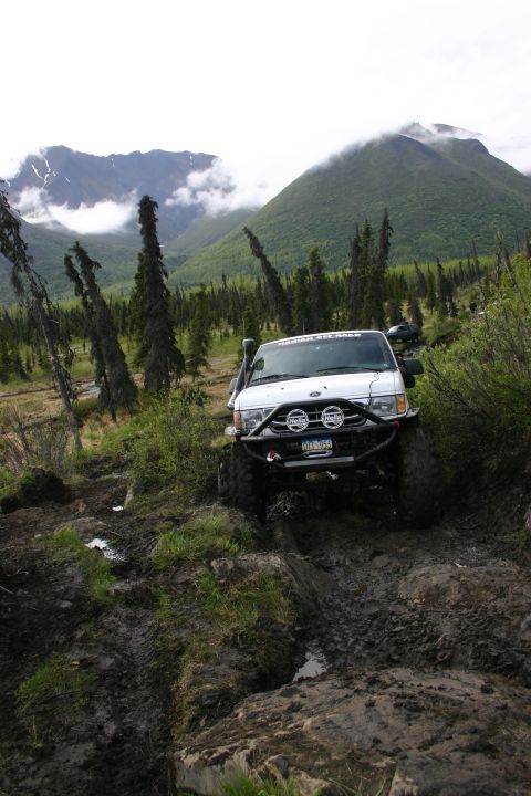 2003 Jeep TJ - A modified van tackles a challenging muddy trail with steep, tree-covered mountains in the distance.