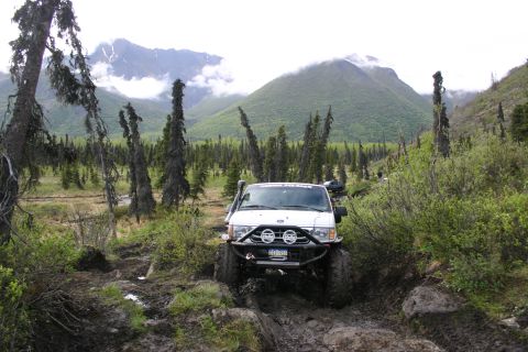 2003 Jeep TJ - A white SUV navigates a challenging muddy path through a wooded area with mountainous terrain ahead.