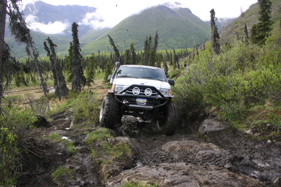 White off-road SUV with snorkel and bull bar navigating a rocky, muddy trail with mountains in the background