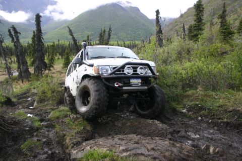 2003 Jeep TJ - A white SUV navigates a challenging muddy off-road course in a mountainous, wooded area.
