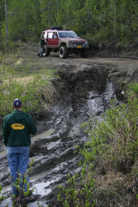 2003 Jeep TJ - Two people attempt to free a Jeep Cherokee XJ from a muddy rut on a wooded trail.
