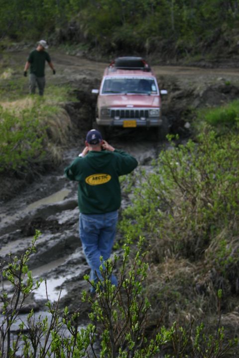 2003 Jeep TJ - A red SUV navigates a muddy trail, with another person visible in the background.