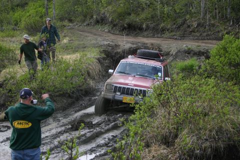 2003 Jeep TJ - A red Jeep Cherokee XJ navigates a challenging muddy trail, with onlookers observing the off-road attempt.