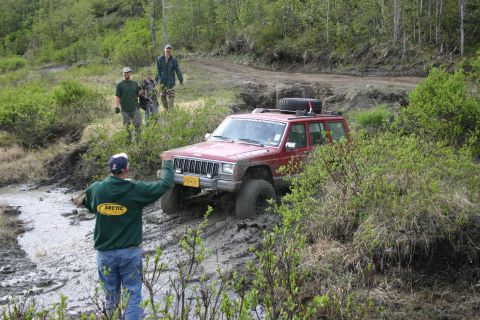 2003 Jeep TJ - A red Jeep Cherokee XJ is stuck in a muddy trail with several people observing the situation.