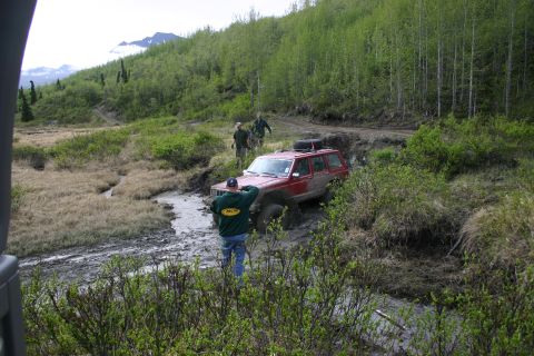 2003 Jeep TJ - A red Jeep Cherokee XJ is stuck in a muddy ditch while two people observe the situation.