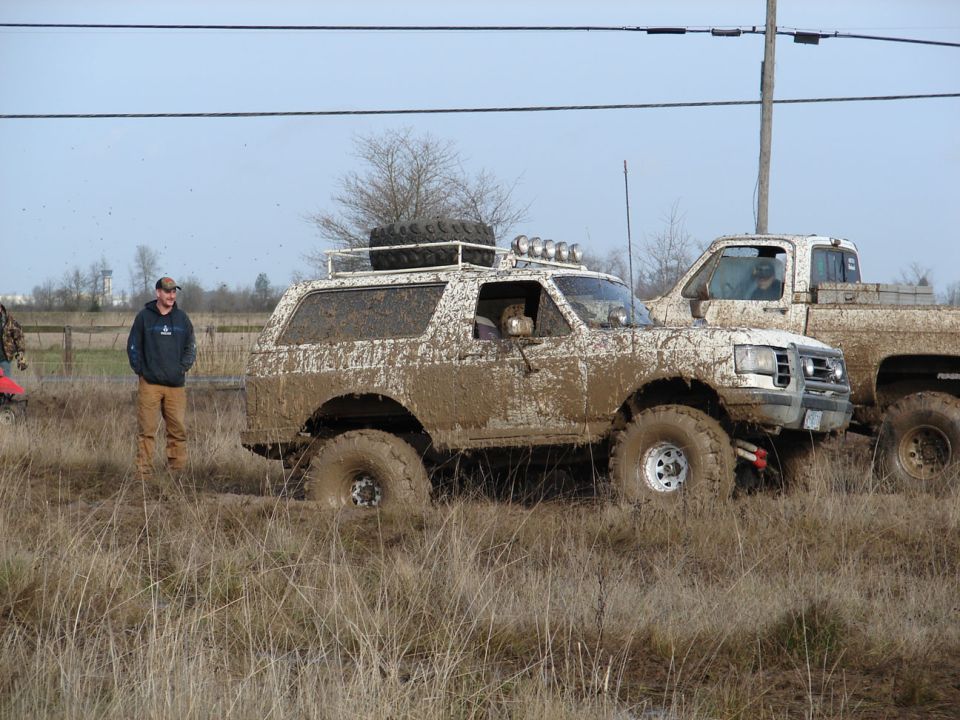 Muddy 1996 Ford Bronco with roof rack and spare tire, parked next to a muddy pickup truck