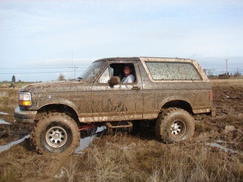 1996 Ford Bronco - A driver navigates a heavily muddied Ford Bronco through a wet, grassy terrain under a cloudy sky.