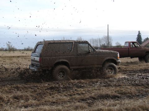 1996 Ford Bronco - A 1996 Ford Bronco navigates a muddy field, kicking up dirt and debris, with another truck visible in the background.