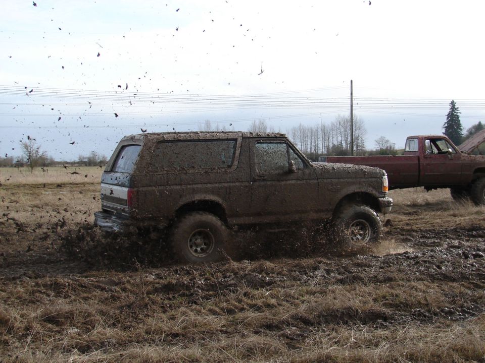 Mud-covered 1996 Ford Bronco splashing through mud with a red pickup truck behind it