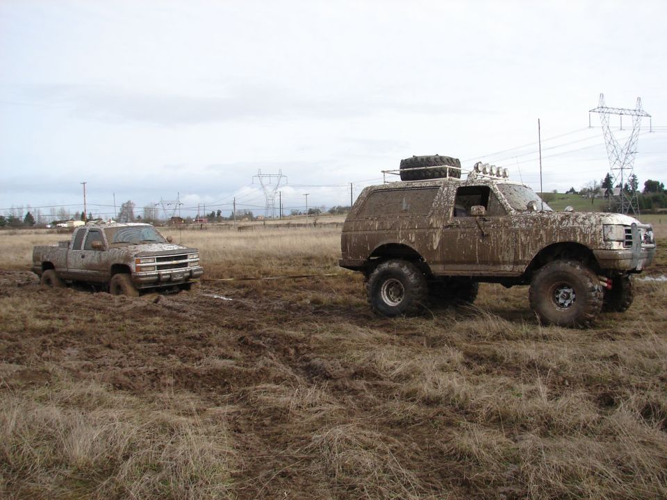 Muddy 1996 Ford Bronco towing a stuck pickup truck in a field