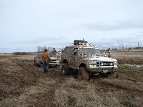1996 Ford Bronco - Two vehicles, a Ford Bronco and a pickup truck, are stuck in a muddy field under an overcast sky.
