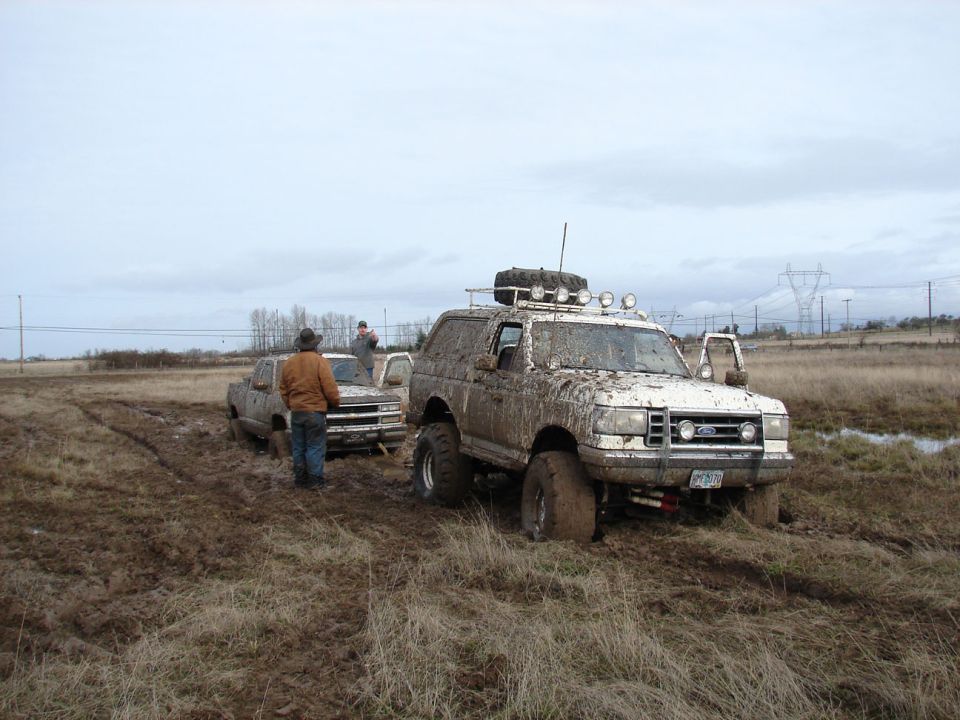 Muddy 1996 Ford Bronco with roof rack and spare tire, stuck in a field next to a pickup truck