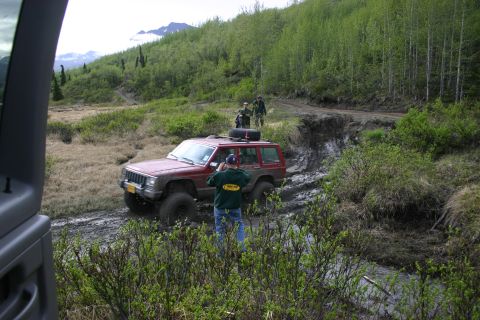 2003 Jeep TJ - A red Jeep Cherokee XJ tackles a challenging muddy off-road path in a wooded area.