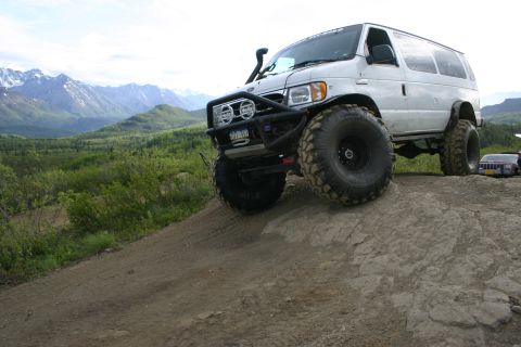 2003 Jeep TJ - A modified white van with extreme off-road tires navigates a rocky trail with a mountainous landscape behind it.