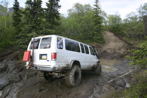 2003 Jeep TJ - A white van equipped with oversized tires tackles a challenging muddy off-road path, with a red gas can attached to the rear.