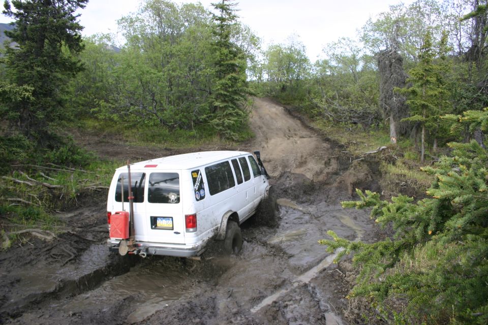 White van struggling on muddy trail, gas can and shovel mounted on back