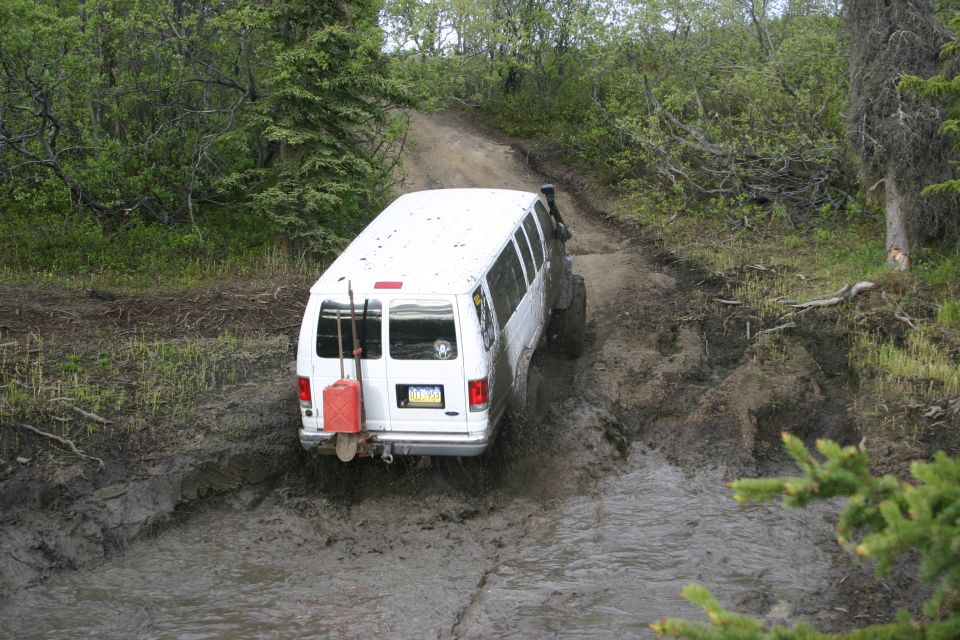 White van struggling on a muddy trail, with a red gas can and shovels mounted on the back