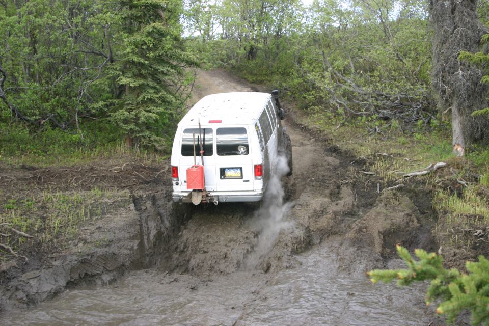White van driving through mud, with a red gas can and shovel mounted on the back