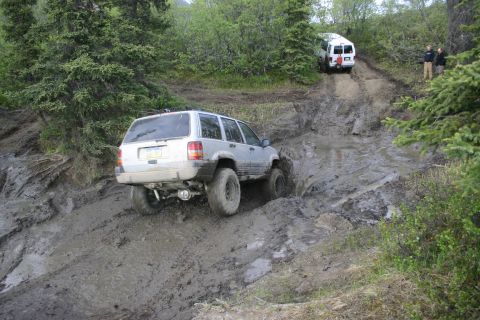 2003 Jeep TJ - A silver 2003 Jeep Grand Cherokee tackles a challenging muddy off-road course, with a white van following closely behind.