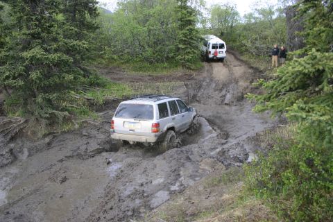 2003 Jeep TJ - Two vehicles navigate a challenging muddy off-road trail with dense trees on either side.