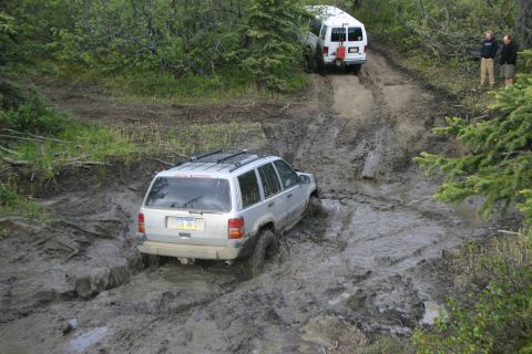 2003 Jeep TJ - A silver SUV struggles through thick mud on an off-road trail, with a white van ahead.