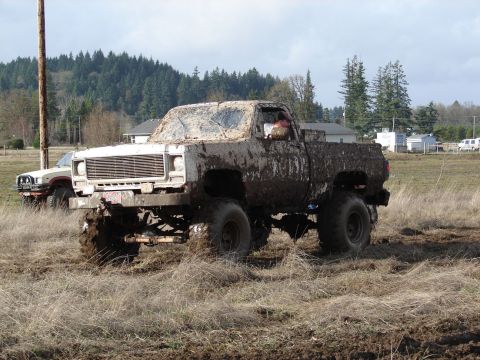 1996 Ford Bronco - A heavily mud-caked pickup truck navigates a muddy field, with another vehicle visible in the background.