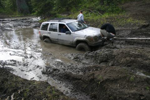 2003 Jeep TJ - A Jeep Grand Cherokee is deeply mired in thick mud, with two individuals attempting to free it.