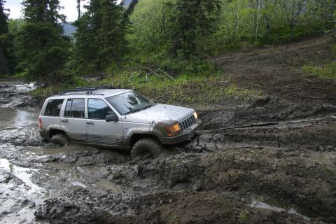 2003 Jeep TJ - A silver SUV is stuck in a muddy trail with trees and foliage in the background.