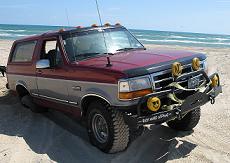 1995 Ford Bronco - This 1995 Ford Bronco is parked on a sandy beach near the ocean, equipped with auxiliary lights and off-road modifications.