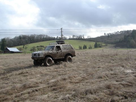1996 Ford Bronco - A heavily muddied Ford Bronco navigates a dry, grassy field under an overcast sky.