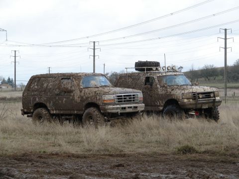 1996 Ford Bronco - Two muddy Ford Broncos, one a 1996 model, sit in a grassy field under an overcast sky.