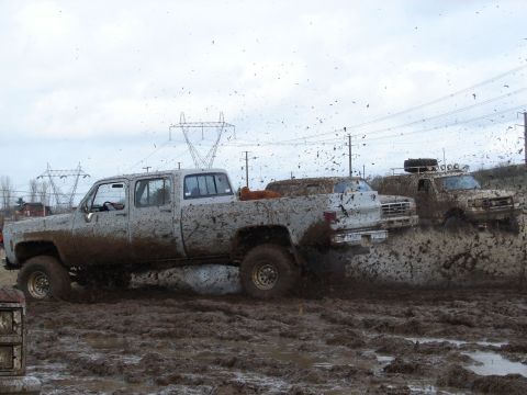 1996 Ford Bronco - Multiple off-road vehicles navigate a challenging muddy terrain, kicking up significant spray.