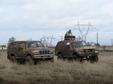 1996 Ford Bronco - Two SUVs are covered in mud after an off-road event, with one vehicle a person on its roof.