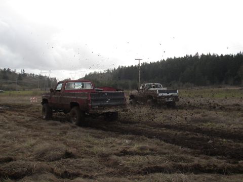 1996 Ford Bronco - Two pickup trucks are driving through a muddy field, kicking up dirt and debris.