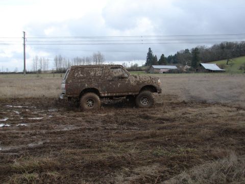 1996 Ford Bronco - A 1996 Ford Bronco is covered in mud while driving through a wet, grassy field with farm buildings in the background.