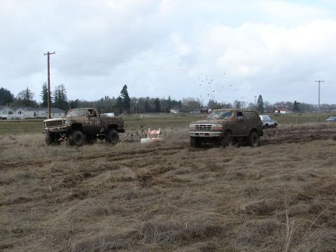 1996 Ford Bronco - Two off-road vehicles navigate a muddy field under an overcast sky.