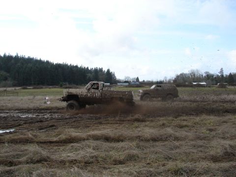 1996 Ford Bronco - Two off-road vehicles are covered in mud as they drive through a wet, grassy field under a cloudy sky.