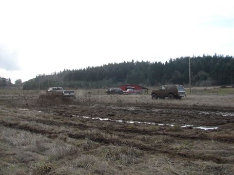 1996 Ford Bronco - A 1996 Ford Bronco navigates a muddy field, kicking up dirt as another truck drives in the distance.