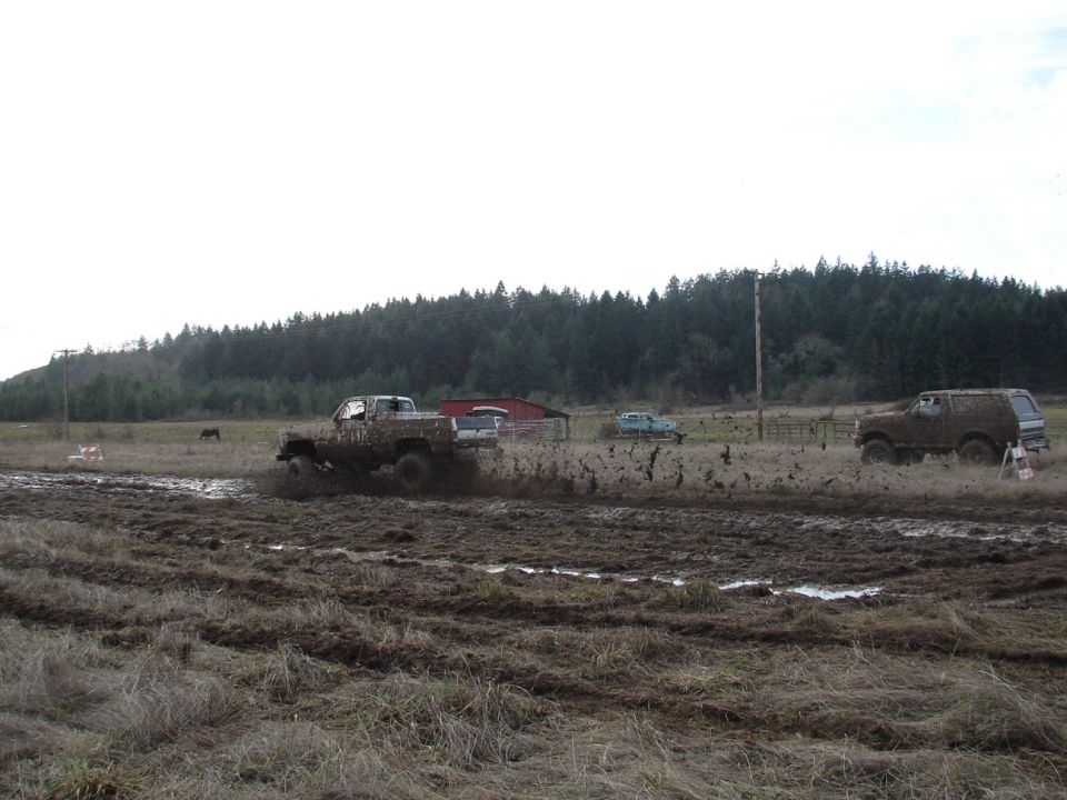 Muddy truck kicking up spray on the left, brown SUV on the right, in a field with a barn and trees