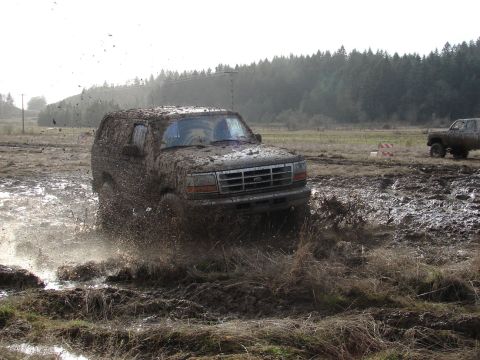 1996 Ford Bronco - This SUV is covered in mud as it drives through a muddy field, with another vehicle visible in the distance.